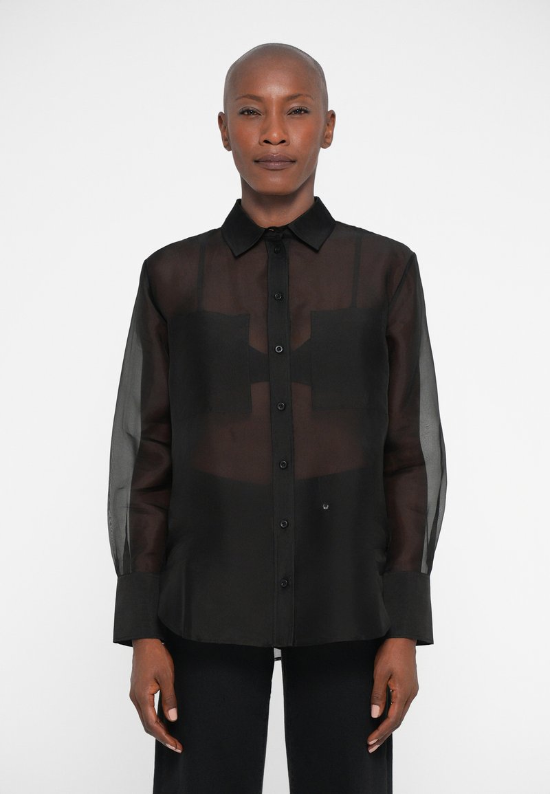 Férfi a kopasz nő, aki fekete áttetsző, hosszú ujjú gombos inget visel egy fekete crop top felett, fehér háttér előtt áll.