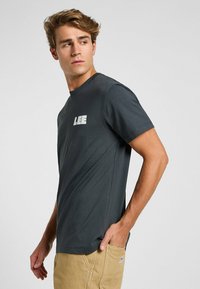 Mörkgrå bomullströja med korta ärmar, prydd med en framträdande vit "LEE"-logotyp på vänster bröst. Slät textur, normal passform.