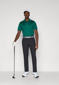 Zelená polokošeľa, sivý opasok, čierne nohavice, biele topánky so sivými akcentmi; golfista drží palicu, má na sebe bielu čiapku a rukavicu.