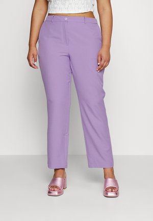 Kobieta w lawendowych, prostych spodniach z wysokim stanem, w parze z metalicznymi różowymi sandałami na słupku z odkrytymi palcami na jednolitym tle.