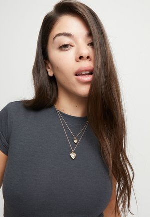 Mujer joven con cabello largo y castaño que lleva collares de colgantes superpuestos en forma de corazón y una camiseta gris oscuro de cuello redondo, sobre un fondo liso.