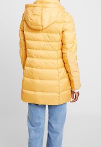 Back view of person wearing a yellow padded coat with hood and light blue jeans, standing against a plain white background.