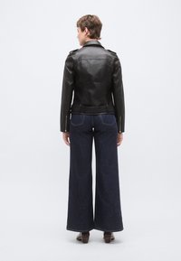 Μαύρο δερμάτινο μπουφάν μοτοσυκλέτας με επισημαντικά, εφαρμοστή γραμμή και πλευρικά φερμουάρ, συνδυασμένο με φαρδιά παντελόνια σκούρου τζιν.