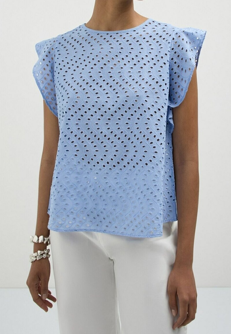 Personne portant un chemisier sans manches bleu clair à motifs ajourés avec des épaules volantées et un pantalon blanc, debout devant un fond uni.