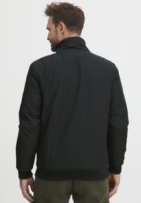 Črna podložena jakna z visokim ovratnikom, rebrastimi manžetami in pasom. Gladka tekstura z minimalističnim dizajnom. Primerna za hladno vreme.