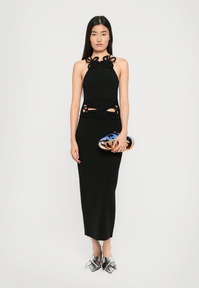 Robe noire à col halter avec des découpes décoratives, une silhouette cintrée, et des accents floraux superposés au niveau du col et de la taille. Pochette argent.