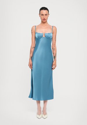 Een blauw satijnen midi-jurk met gegolfde boezemaccenten, dunne schouderbandjes en een zijsplit, gecombineerd met witte spitse hakken.