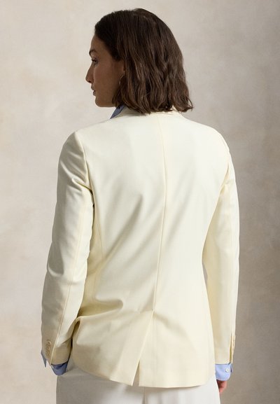 Vállig érő barna hajú nő törtfehér blézert visel világoskék ing felett, hátulnézetben, semleges háttér előtt.