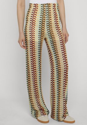 Frau trägt hoch taillierte, weit geschnittene Hosen mit mehrfarbigen vertikalen wellenförmigen Streifen in Rot, Blau, Beige und Braun, kombiniert mit weißen Turnschuhen.