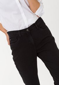 Jeans de mezclilla negra con un diseño ajustado, que cuentan con bolsillos frontales, un cierre de botón y una textura suave. Combinados con una camisa blanca de botones.