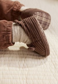 Chaussons en velours côtelé marron avec une texture côtelée et un motif à carreaux sur les semelles, portés avec des chaussettes gris clair, posés sur une surface matelassée crème.