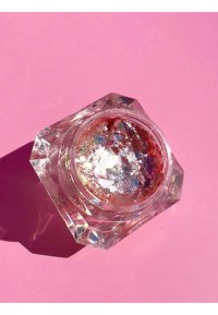 Barattolo esagonale trasparente contenente scaglie glitterate e iridescenti con sfumature di rosso. Il barattolo è posato su uno sfondo rosa, proiettando un'ombra.
