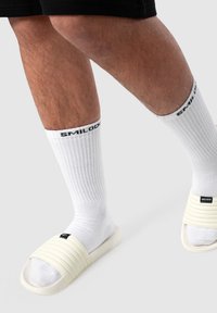 Sandali a scivolo di colore crema con fasce testurizzate e rigate. Indossati sopra calze bianche al ginocchio con marchio nero. Mostrano i piedi e la parte bassa delle gambe.
