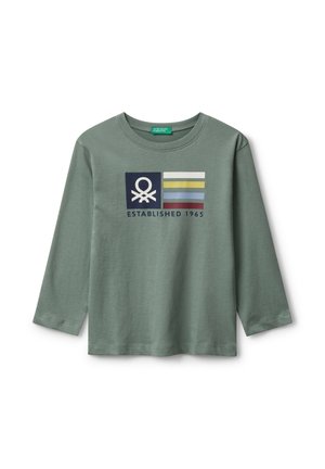 Camicia verde a maniche lunghe con un logo e strisce colorate sopra il testo "Fondata nel 1965" al centro davanti.