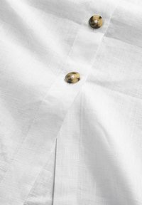 Chemise en lin blanche avec deux boutons marbrés marron et une texture subtile, dotée d'une patte de boutonnage sur le devant.