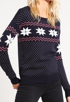 Femme portant un pull bleu marine avec des motifs d'étoiles blanches, des lignes en zigzag roses et de petits points blancs, associé à un pantalon noir.