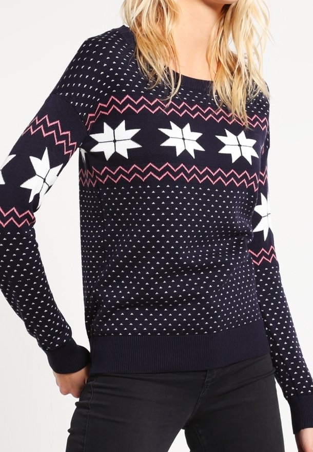 Femme portant un pull bleu marine avec des motifs d'étoiles blanches, des lignes en zigzag roses et de petits points blancs, associé à un pantalon noir.