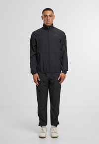 Černá lehká bunda s vysokým límcem, předním zipem a elastickými manžetami, doplněná o odpovídající kalhoty. Hladká textura, minimální branding.