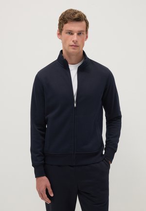 Jonge man met kort krullend haar, gekleed in een marineblauwe jas met rits over een wit shirt, met de handen omlaag en één hand in de zak, tegen een witte achtergrond.