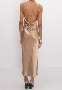 Robe midi en satin beige avec un dos drapé, des bretelles croisées et une texture lisse, associée à des sandales à talons épais.