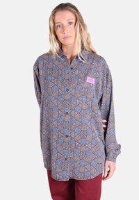 Femme blonde portant une chemise à motifs lâche avec des designs géométriques et un pantalon bordeaux, se tenant avec une main derrière le dos.