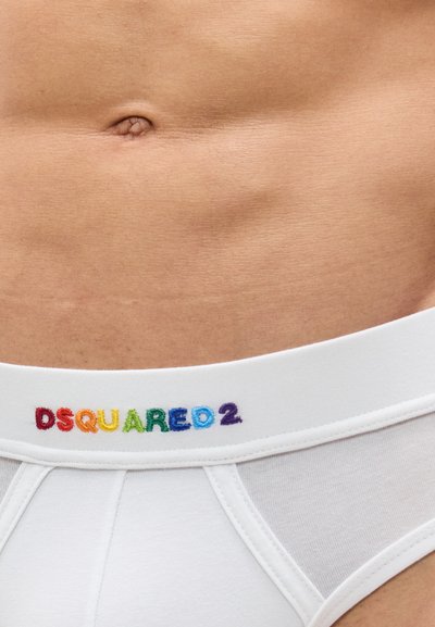 Κοντινή λήψη της κάτω κοιλίας ενός ατόμου που φοράει λευκά εσώρουχα με το ουράνιο τόξο λογότυπο "DSQUARED2" στη ζώνη μέσης.