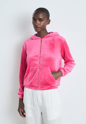 Sweat à capuche zippé en velours rose avec une poche frontale et un cordon de serrage, présentant une texture lisse et une coupe décontractée.