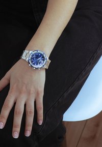 Silver rostfritt stål klocka med en blå urtavla, prydd med en bezel med blå accenter, med en kronografdesign. Bar på ett naket handled.