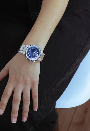 Orologio in acciaio inossidabile argentato con quadrante blu, lunetta decorata con accenti blu e design cronografo. Indossato su un polso nudo.