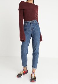 Bordový svetr s odhalenými rameny z pleteniny, džíny s vysokým pasem a vícerozměrné tenisky s puntíkovaným vzorem a kontrastními akcenty.