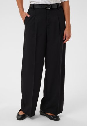 Personne portant un pantalon noir à jambes larges avec une ceinture noire, une chemise blanche et des ballerines noires, debout avec une main dans la poche.