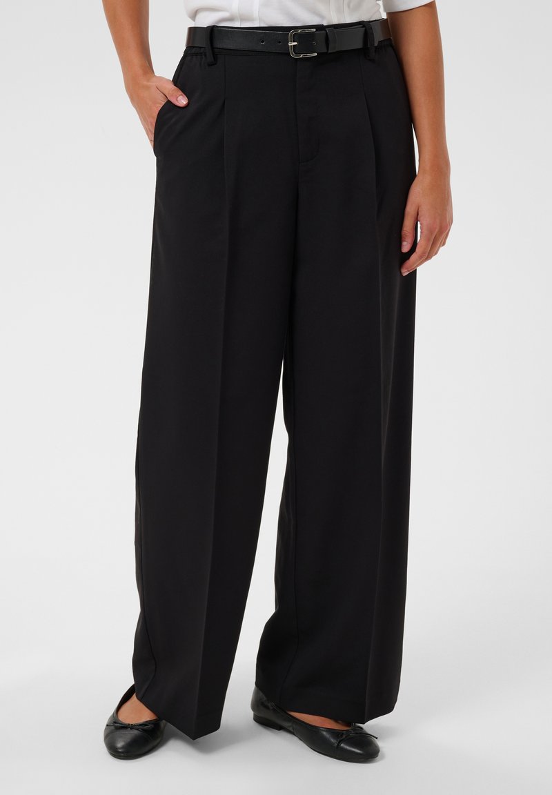 Personne portant un pantalon noir à jambes larges avec une ceinture noire, une chemise blanche et des ballerines noires, debout avec une main dans la poche.