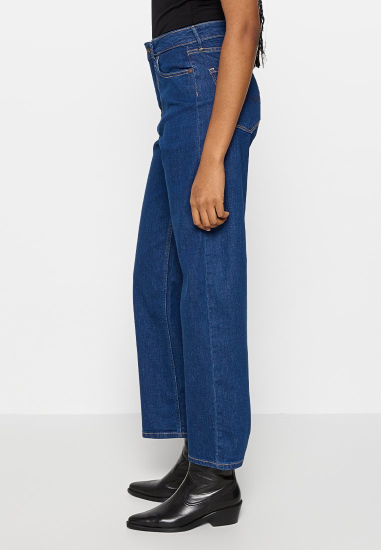 Jean en denim bleu à jambes larges avec taille haute, présentant un design classique à cinq poches et des coutures orange contrastantes.