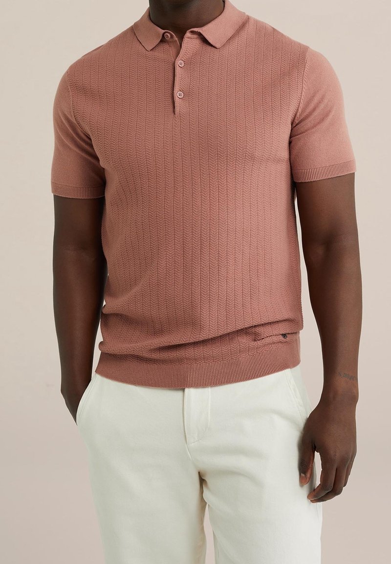 Homme portant un polo à manches courtes texturé rose poudré associé à un pantalon blanc cassé, les mains partiellement dans les poches.
