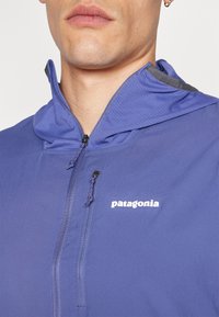 Purpurna jakna s kapuco iz lahkega materiala, ki ima zadrgo, žep na prsih in logo Patagonia spredaj.