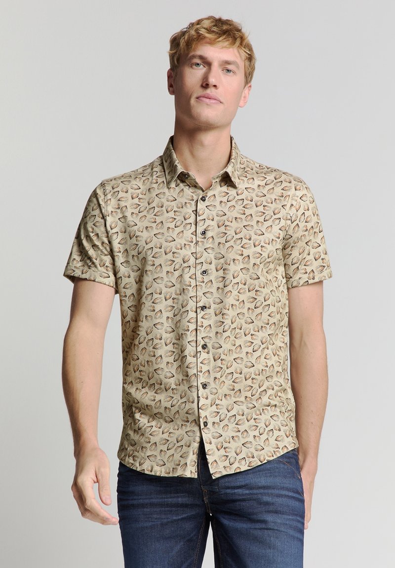 Korte mouwen shirt in lichtbruine stof met een schelp patroon, voorzien van een button-down kraag en zwarte knopen. Gekleed met donkerblauwe jeans.
