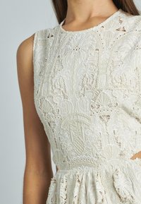 Abito beige chiaro con ricami intricati caratterizzati da motivi floreali e geometrici; include dettagli a taglio in vita e gonna fluida.