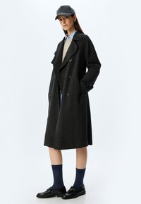 Donkergrijze wollen trenchcoat met grote kraag en dubbele knopen. Gecombineerd met een lichtblauwe blouse, een beige trui en zwarte schoenen.