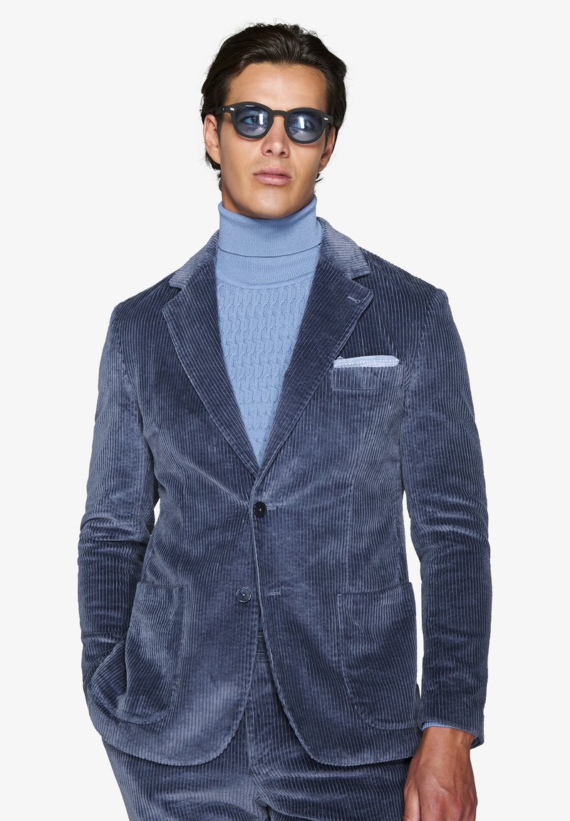Blazer in velluto a coste blu navy, con vestibilità tailored, due tasche frontali, rever a punta, abbinato a un maglione a collo alto blu chiaro.
