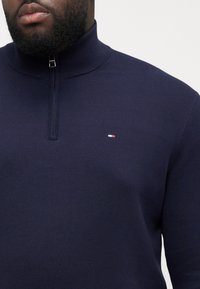 Granatowy sweter z wysokim kołnierzem, zamek błyskawiczny, struktura ściągacza oraz małe logo w kolorach czerwonym, białym i niebieskim na piersi.