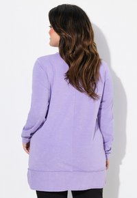 Tunic lunga lavanda con maniche lunghe e orlo arrotondato, in morbido tessuto testurizzato e dettagli di cucitura discreti sulla schiena. Indossata su pantaloni neri.