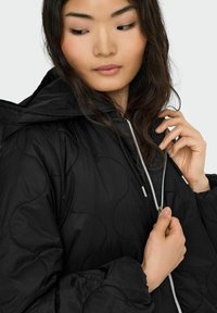 Schwarze gesteppte Jacke mit Kapuze, einem silbernen Reißverschluss und strukturierten Steppnähten. Der Stoff wirkt glatt und leicht.