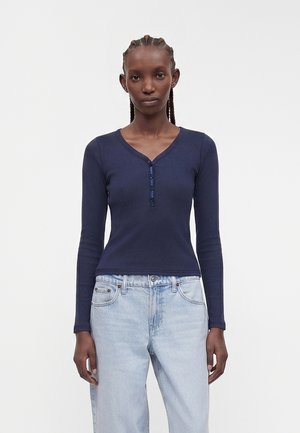 Kobieta z warkoczami, ubrana w granatową bluzkę henley z długim rękawem oraz jasnoniebieskie, wysokie jeansy, stoi na jednolitym tle.