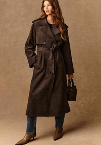 Cappotto trench in suede marrone con vita a cintura, grandi bottoni e colletto classico. Abbinato a jeans blu e stivaletti a punta marroni.