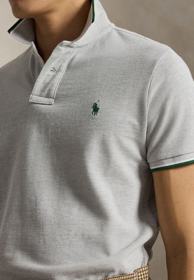 Gros plan d'une personne portant un polo gris clair avec une bordure verte au col et à la manche, avec un petit logo vert brodé.