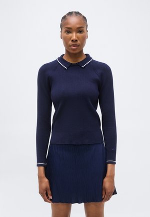 Kvinde med flettet hår iført en marineblå langærmet polotrøje og et plisseret navyblåt skørt mod en ensfarvet lys baggrund.