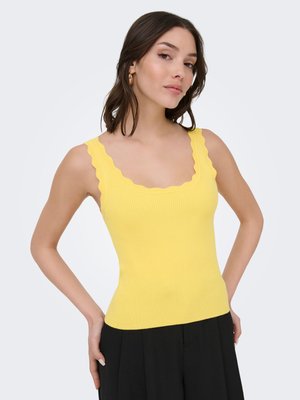 Femme aux cheveux mi-longs foncés, portant des boucles d'oreilles créoles dorées, un haut sans manches jaune à bord festonné et un pantalon noir, les mains sur les hanches.