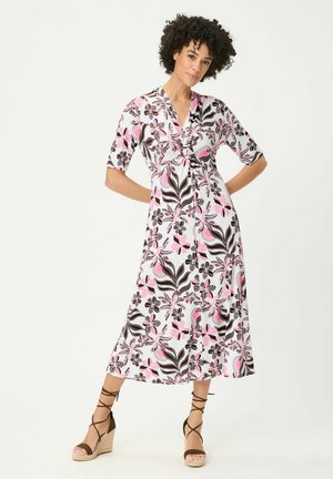 Frau trägt ein weißes Midikleid mit pinkem und schwarzem Blumenmuster, Ellbogenlange Ärmel und braune Keilsandalen mit Knöchelriemen.