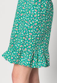 Saia floral verde com bainha franzida, feita de um tecido leve com padrões de flores em rosa, branco e amarelo por toda a parte.