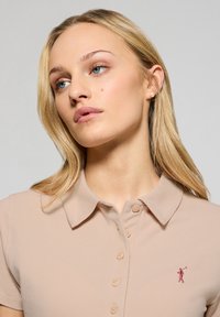 Beige polo shirt med korta ärmar, knapplängd fram, och en subtil broderad design av en golfspelare. Mjuk textur med en klassisk krage.